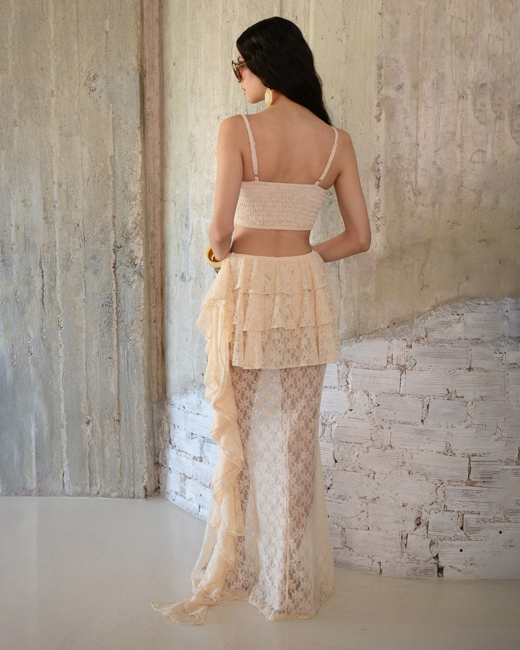 Woman wearing a beige top and lace skirt against a textured wall.
