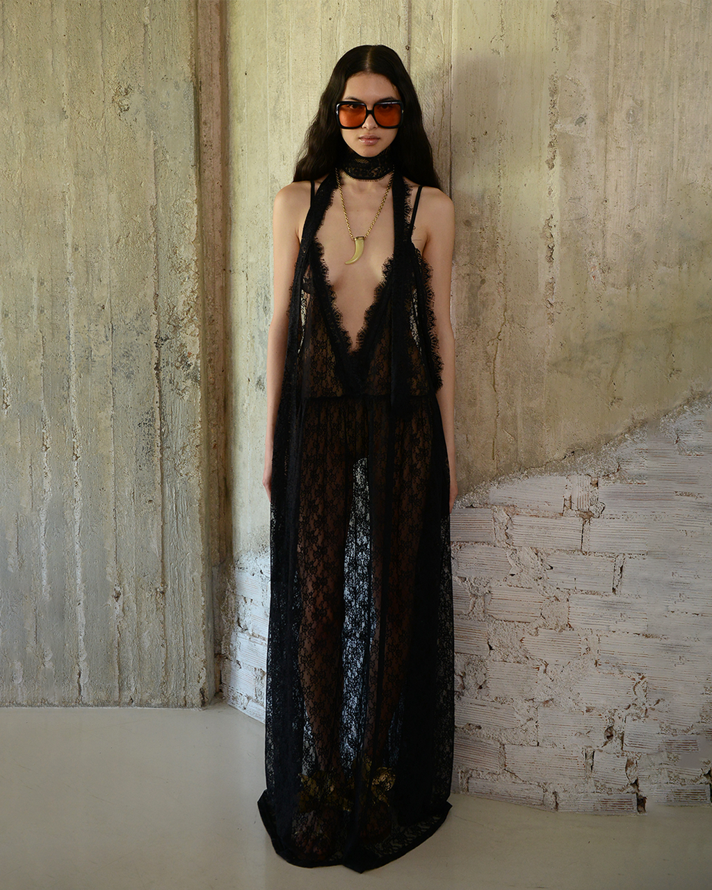 Woman wearing a black lace dress and sunglasses against a textured wall.