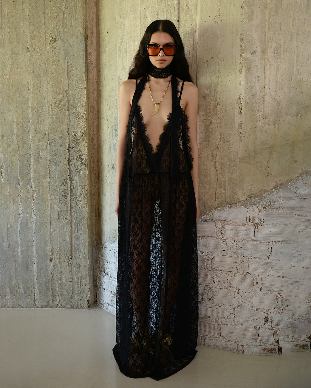 Woman wearing a black lace dress and sunglasses against a textured wall.