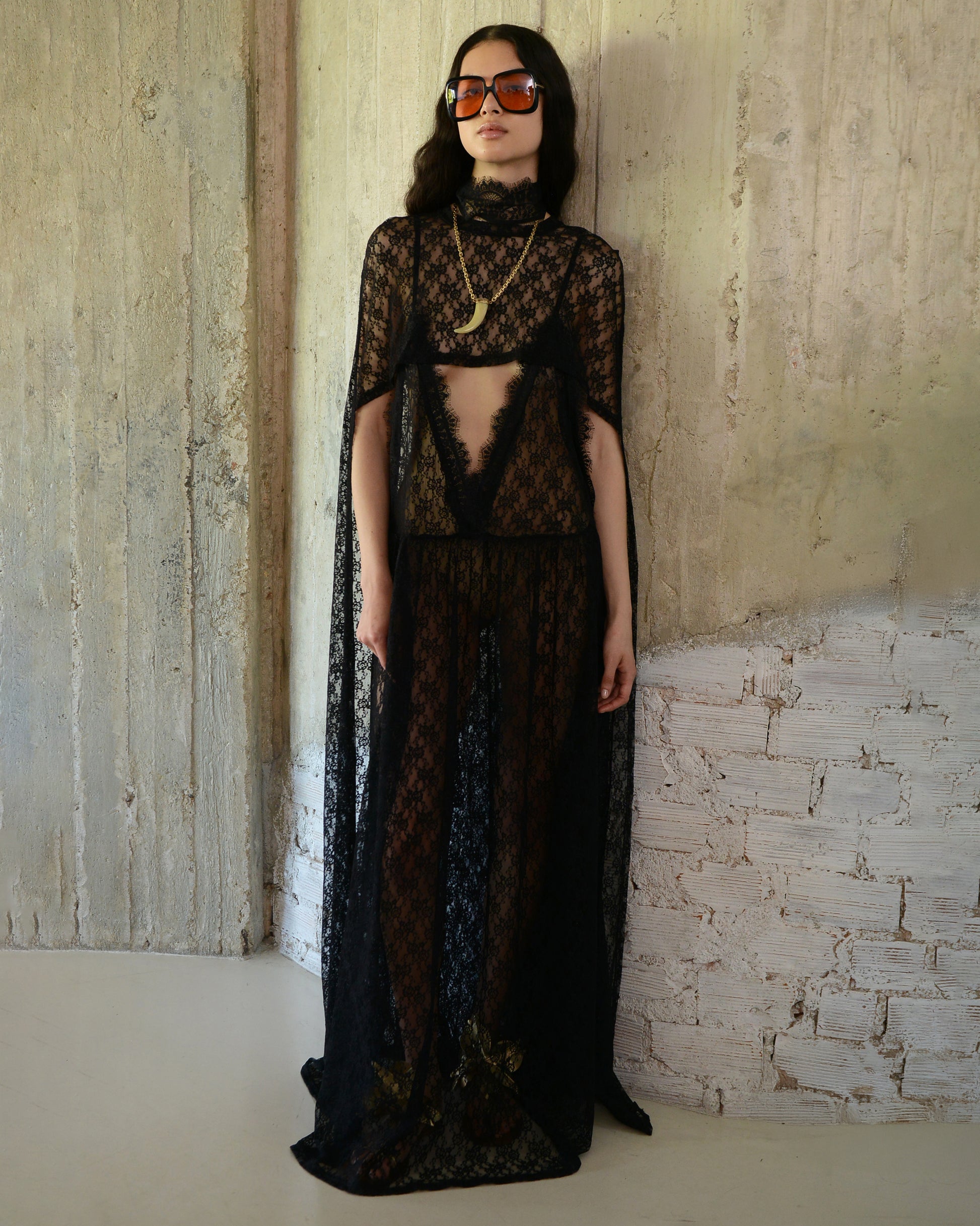 Woman wearing a black lace dress with a cape against a textured wall.