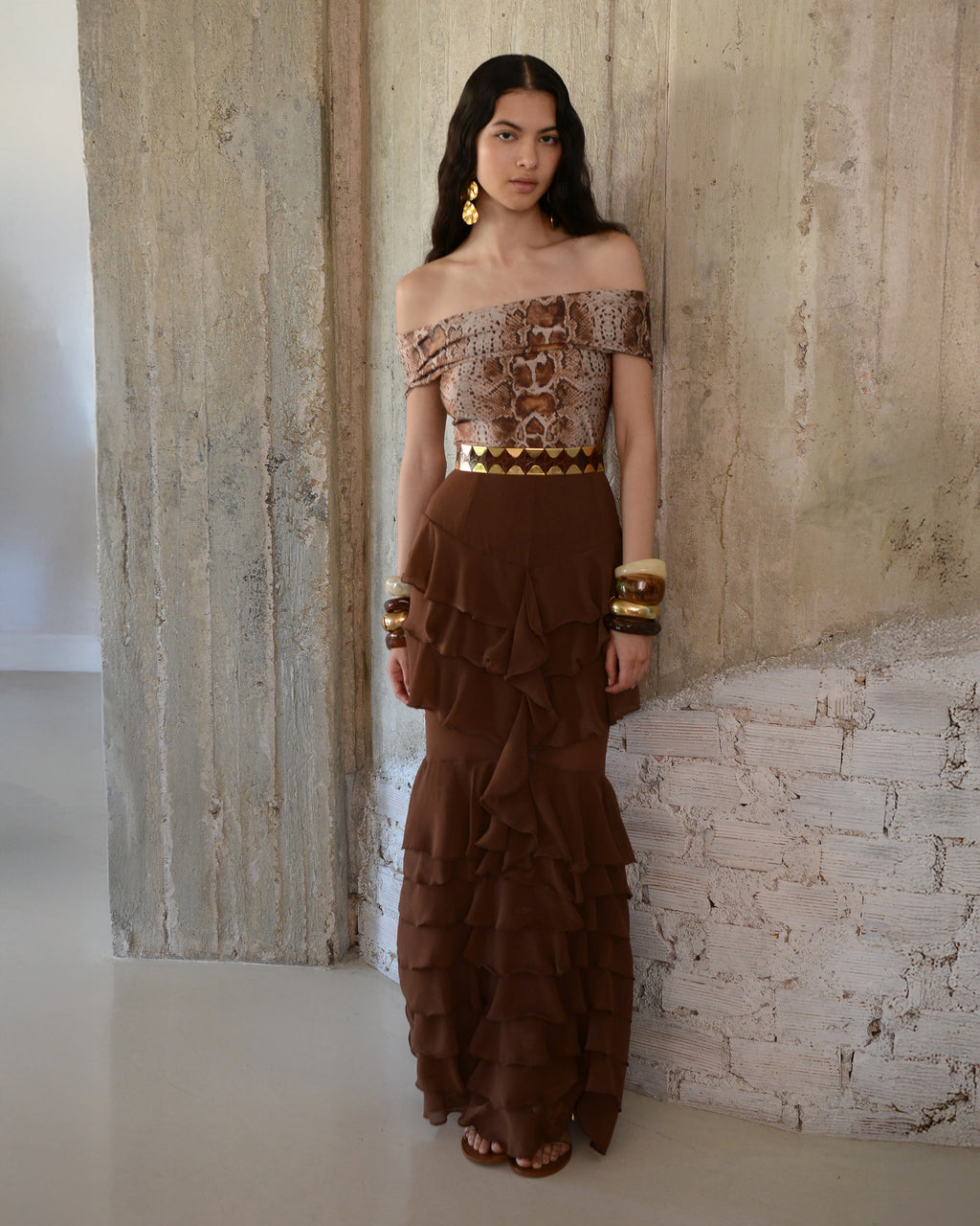 Woman wearing a brown off-shoulder top and ruffled skirt against a textured wall.