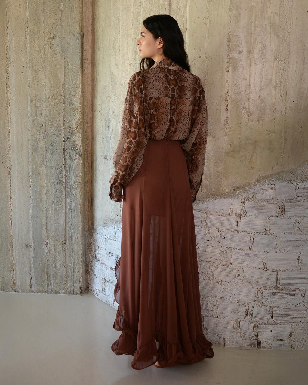 Woman wearing a brown outfit with a patterned top and long skirt against a textured wall.