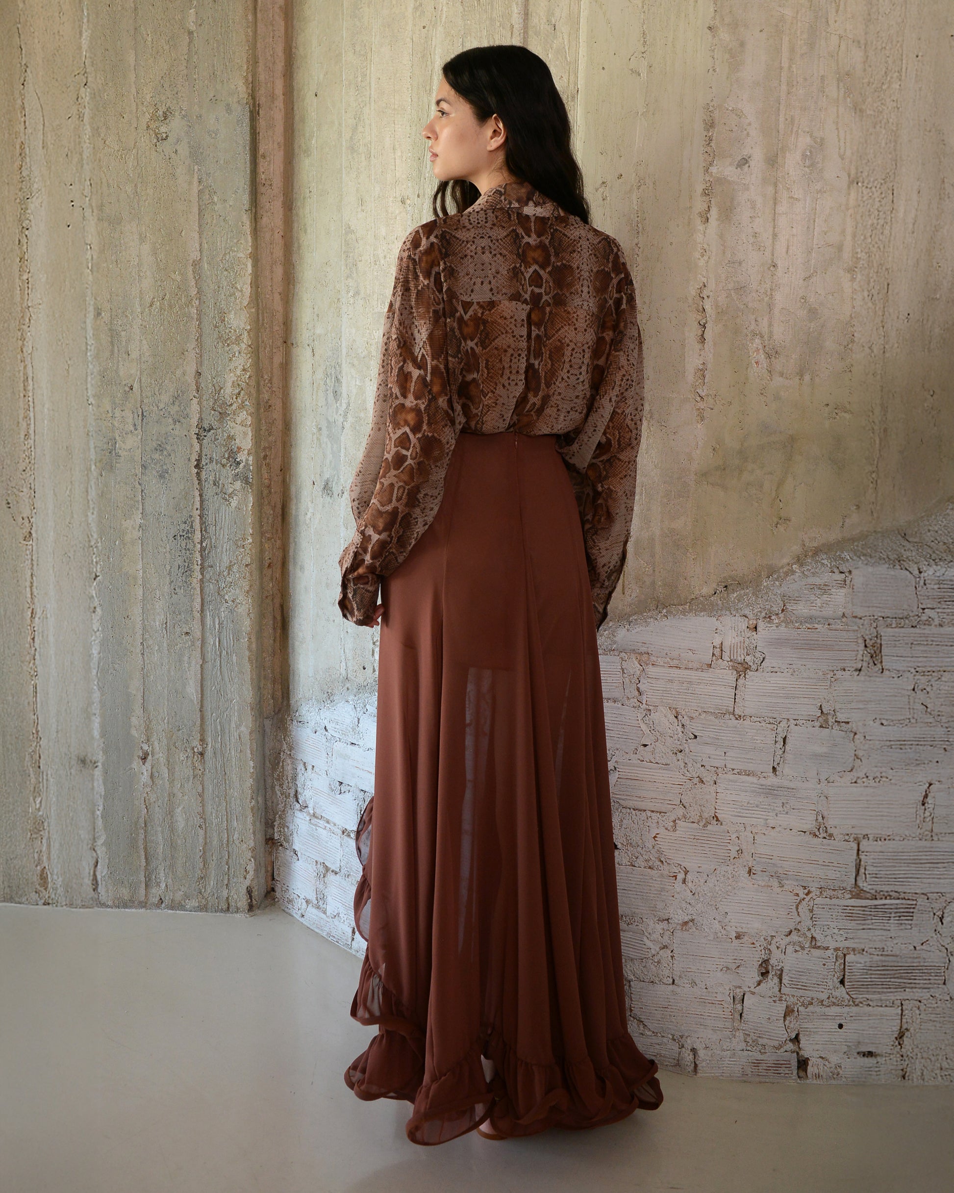 Woman wearing a brown outfit with a patterned top and long skirt against a textured wall.