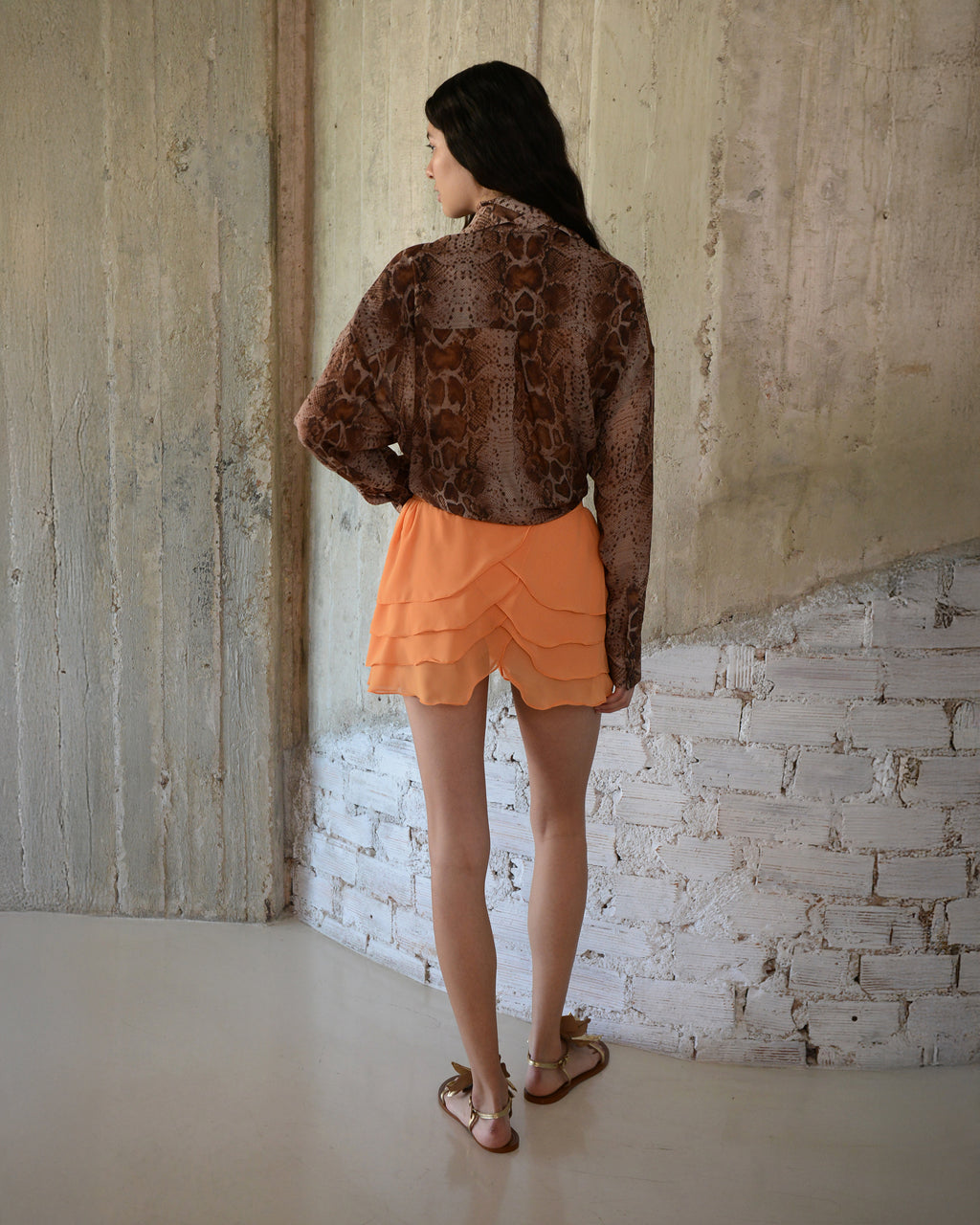 Woman wearing a brown patterned top and orange shorts standing against a textured wall.