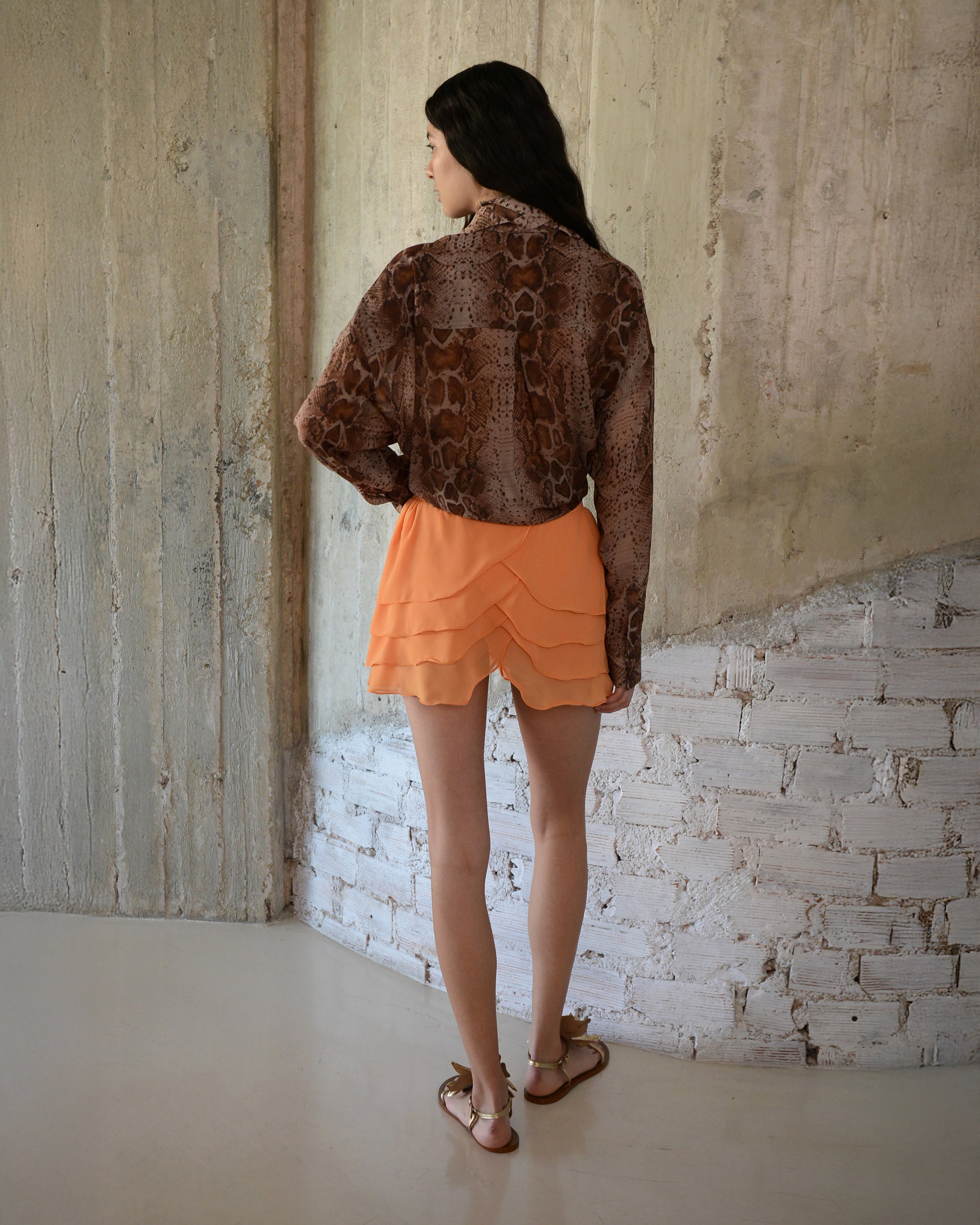 Woman wearing a brown patterned top and orange shorts standing against a textured wall.