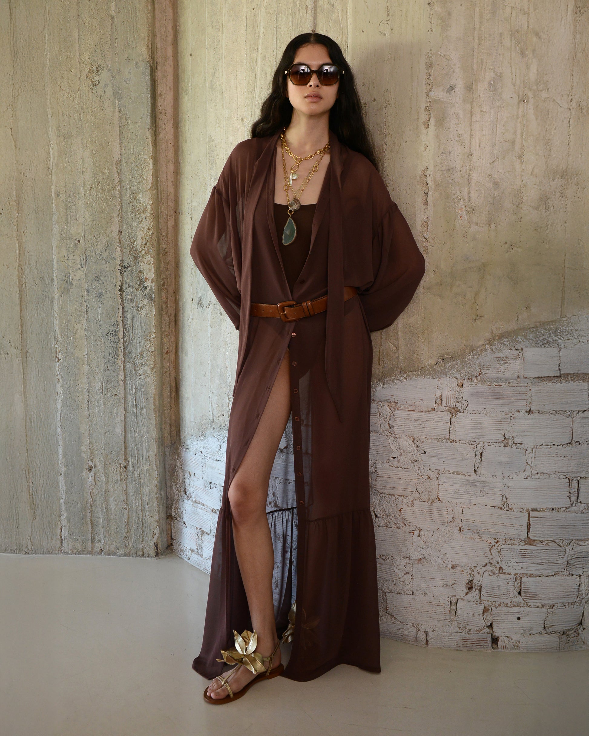 Woman wearing a long brown dress with a slit, standing against a textured wall.