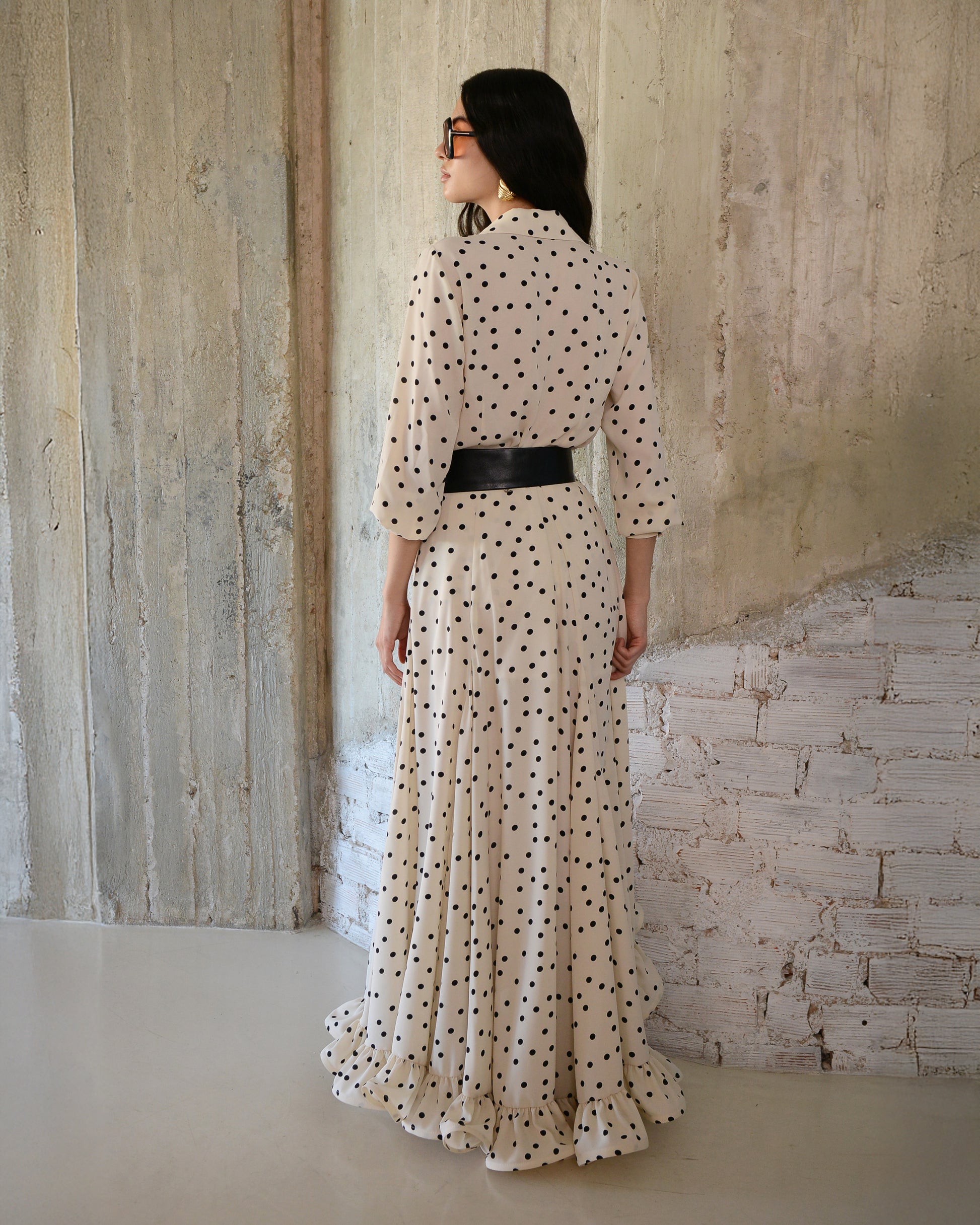Woman wearing a long, light-colored dress with black polka dots against a textured wall.