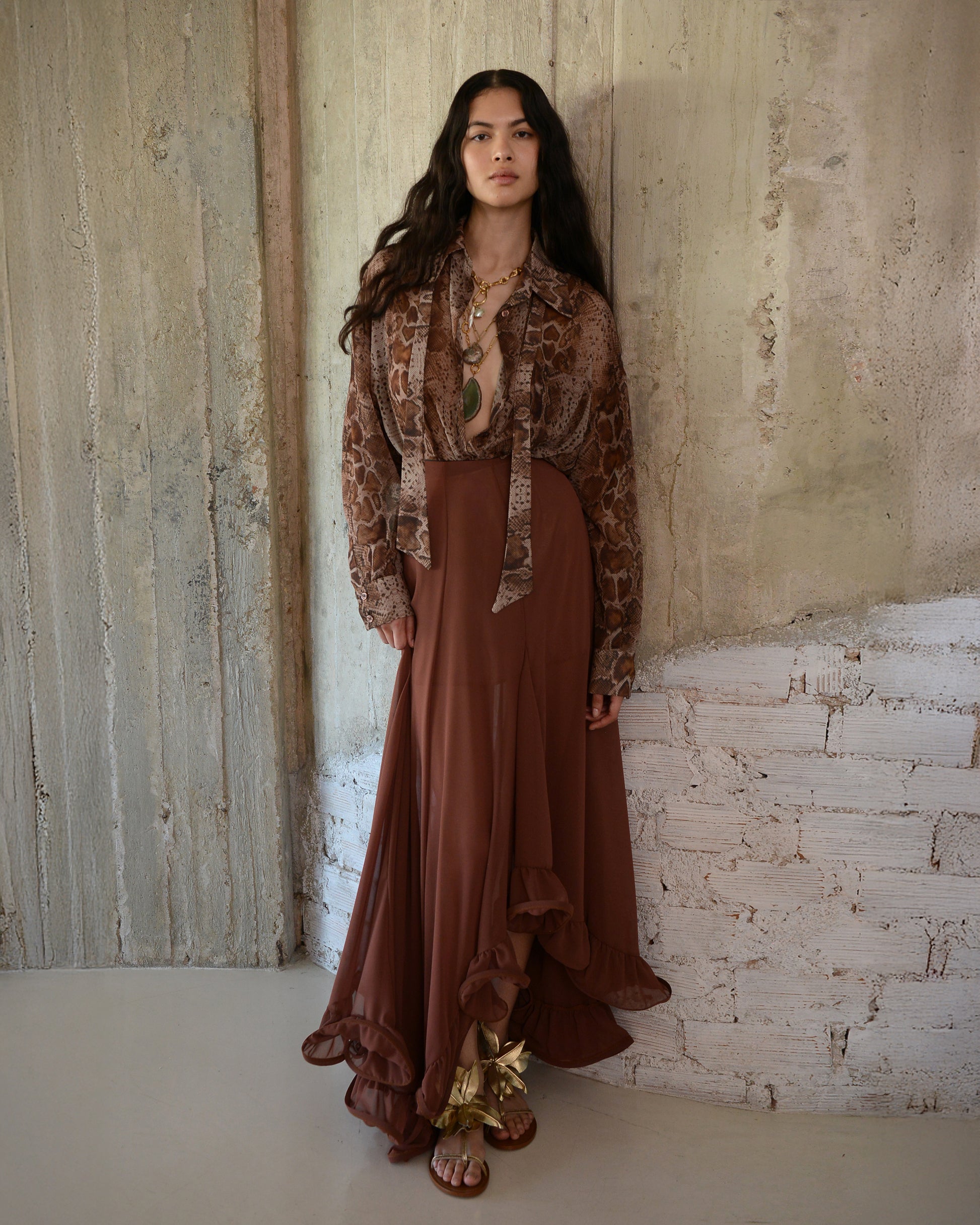 Woman wearing a patterned top and brown dress standing against a textured wall.