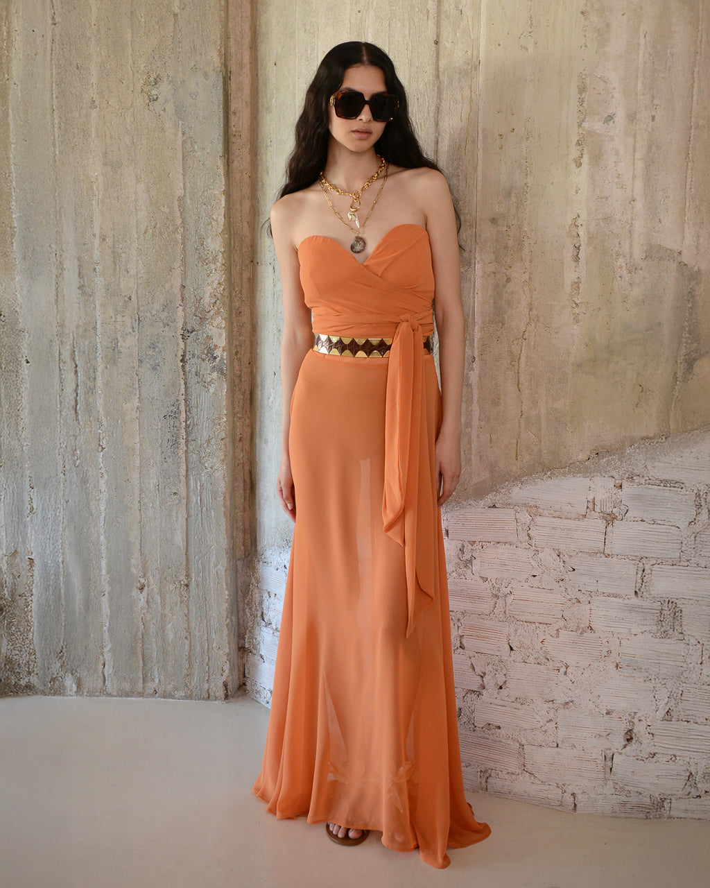 Woman wearing an orange strapless dress with a belt against a textured wall.