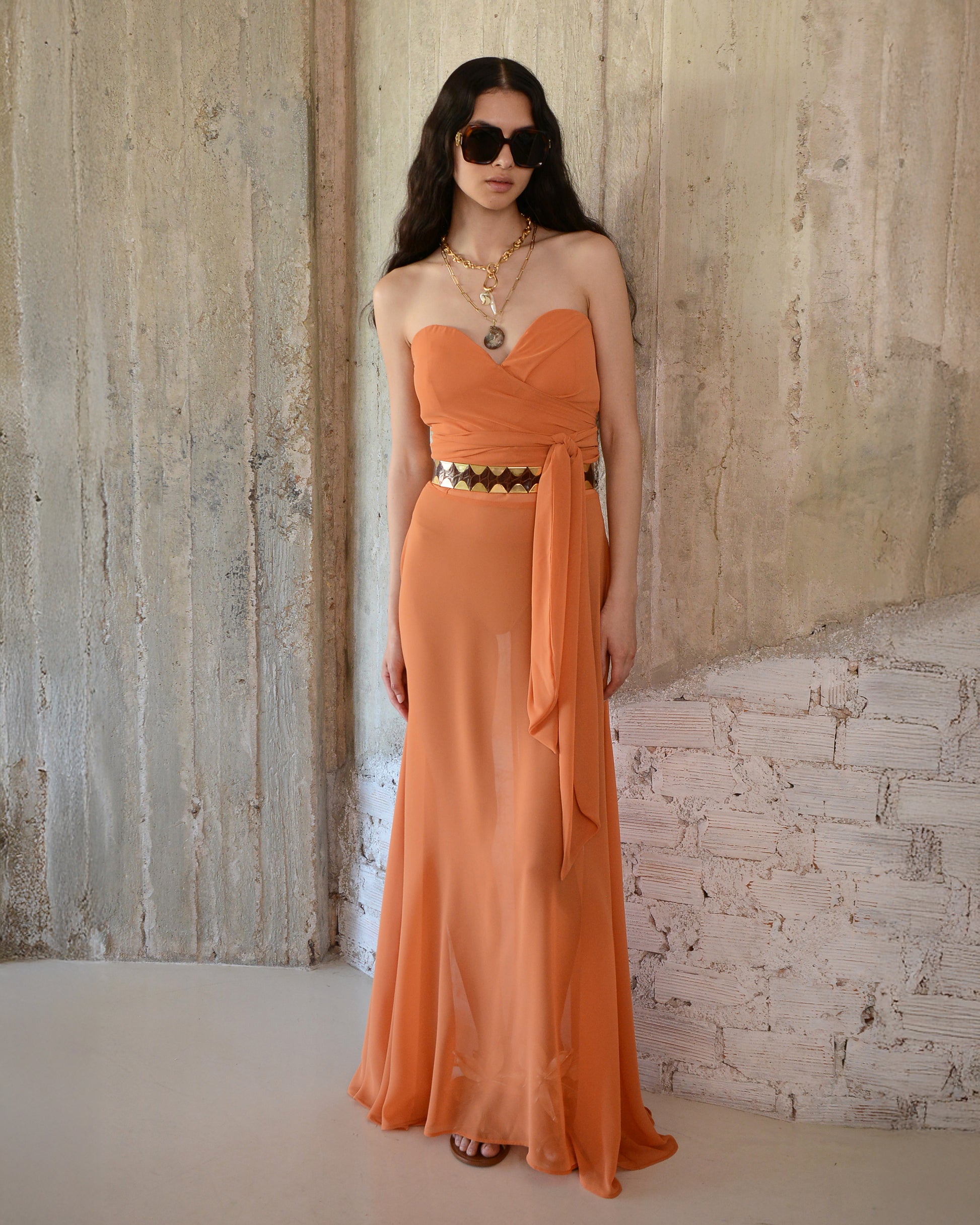 Woman wearing an orange strapless dress with a belt against a textured wall.