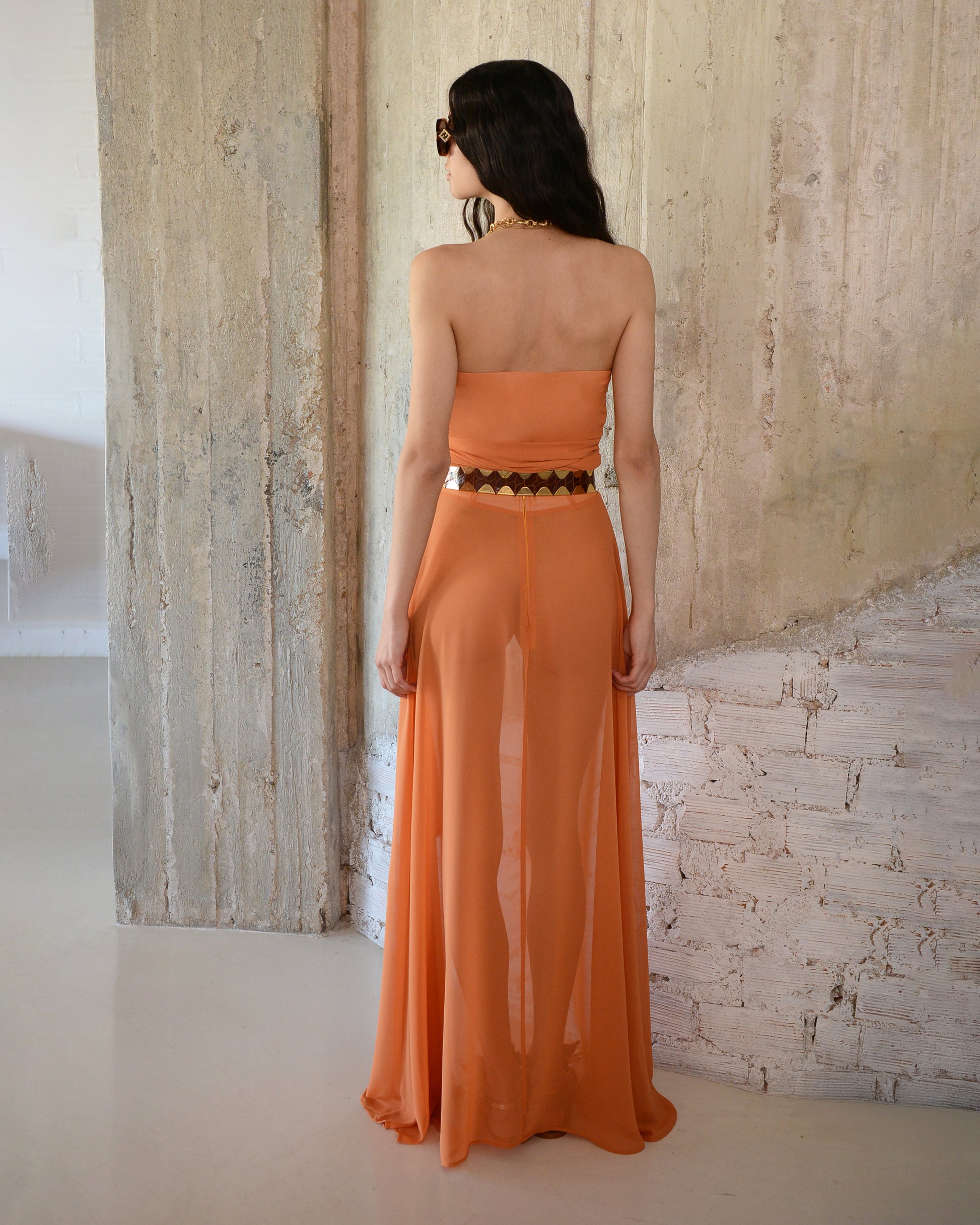 Woman wearing an orange strapless dress with a geometric belt against a textured wall.